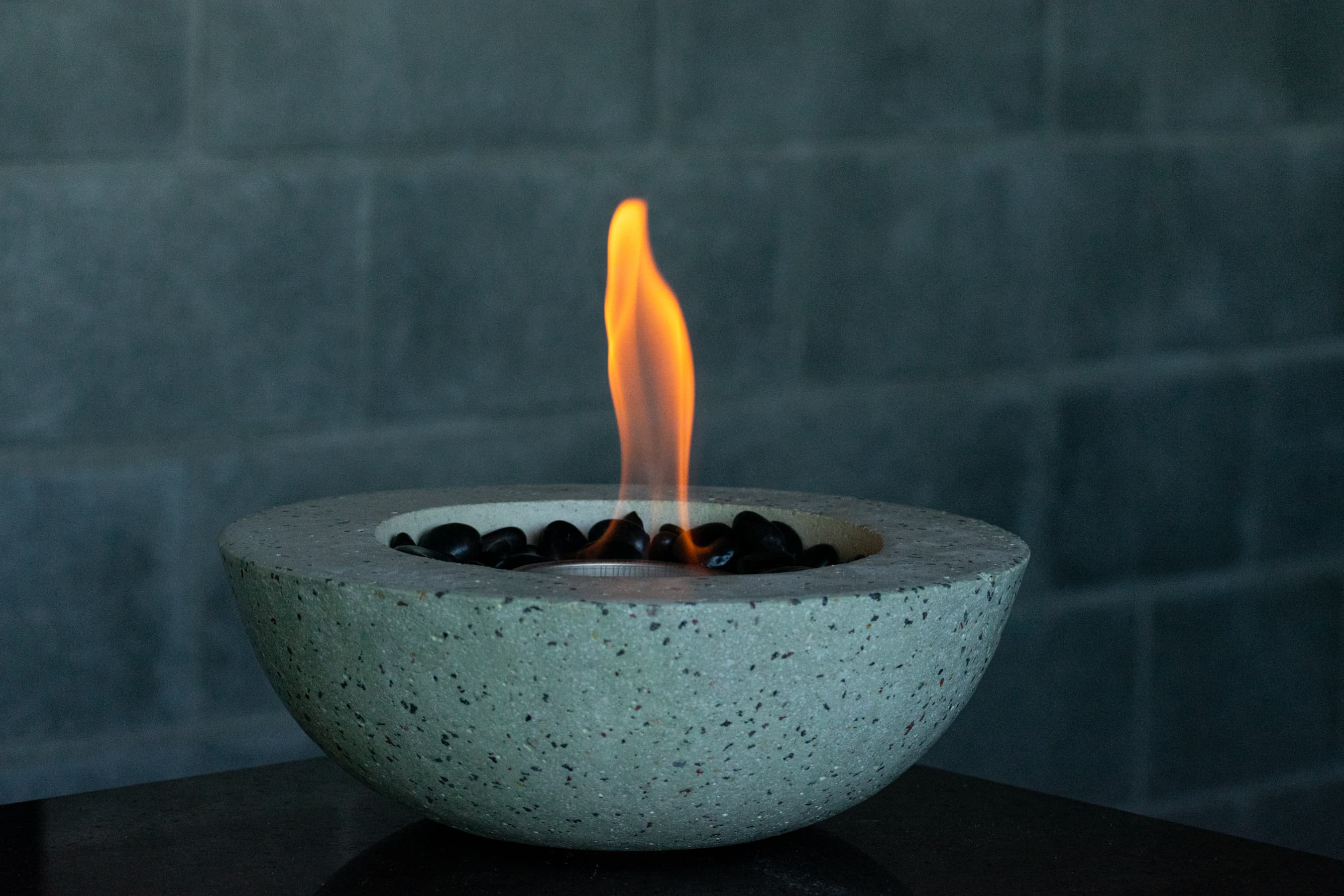 Fuego de mesa de concreto encendido, con piedras negras, frente a una pared de bloques
